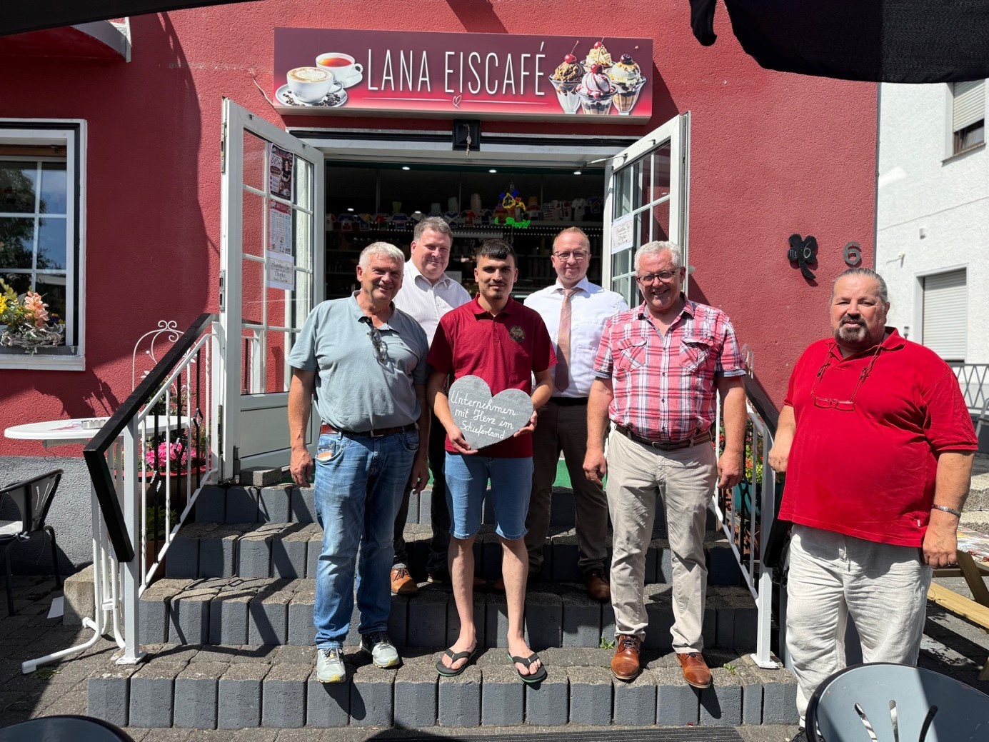 Die Aufnahme zeigt sechs Männer vor einem roten Gebäude, das als "Lana Eiscafé" ausgeschildert ist. Sie stehen auf einer Steintreppe, die zur Eingangstür führt. Die Sonne scheint, was zu deutlichen Schattenwürfen führt. Im Hintergrund ist die Fassade des Eiscafés in einem kräftigen Rotton gehalten. Über dem Eingang hängt ein Schild mit dem Namen des Cafés in weißer Schrift, daneben Bilder von Kaffee und Eisbechern. Die Eingangstür und die beiden angrenzenden Fenster sind aus Glas und weiß. Links neben der Tür befindet sich ein Fenster mit Blumenkästen, die mit rosa Blumen und etwas Grünzeug geschmückt sind. Auf dem Sims davor steht ein kleiner weißer Tisch. Die sechs Männer sind in unterschiedlichen Altersgruppen und in unterschiedlicher Kleidung. Alle lächeln und scheinen posiert zu sein. Von links nach rechts: Der erste Mann von links hat graue Haare, trägt ein hellblaues Polohemd und Jeans. Seine Sonnenbrille hängt über dem Kragen. Der zweite Mann ist etwas jünger, trägt ein weißes Hemd und hat dunkle Haare. Er steht etwas im Schatten. Der dritte Mann, der jüngste in der Gruppe, trägt ein dunkelrotes Poloshirt und blaue Shorts. Er hält ein herzförmiges Schild mit deutscher Aufschrift "Unternehmern mit Herz im Sauerland" in den Händen. Er trägt Flip-Flops. Der vierte Mann hat graue Haare, trägt eine Brille, ein weißes Hemd mit einer leicht geöffneten Krawatte und eine dunkelbraune Hose. Der fünfte Mann von links ist deutlich älter, trägt ein kariertes Hemd in Rot- und Weißtönen, beige Hosen und braune Schuhe. Der sechste Mann, ganz rechts im Bild, hat kräftige Statur, kurze dunkle Haare und einen Bart. Er trägt ein knallrotes Poloshirt und helle Hosen. Seine Sonnenbrille trägt er auf dem Kopf. Die Beleuchtung ist hell und frontal, was die Farben des Gebäudes und der Kleidung hervorhebt. Die Komposition ist mittig, die Männer sind auf einer Linie arrangiert, was eine freundliche und einladende Atmosphäre schafft. Die grauen Steinstufen der Treppe bilden einen klaren Vordergrund und eine deutliche Trennung zum Hintergrund. Die Schatten auf den Stufen und den Männern geben dem Bild Tiefe.