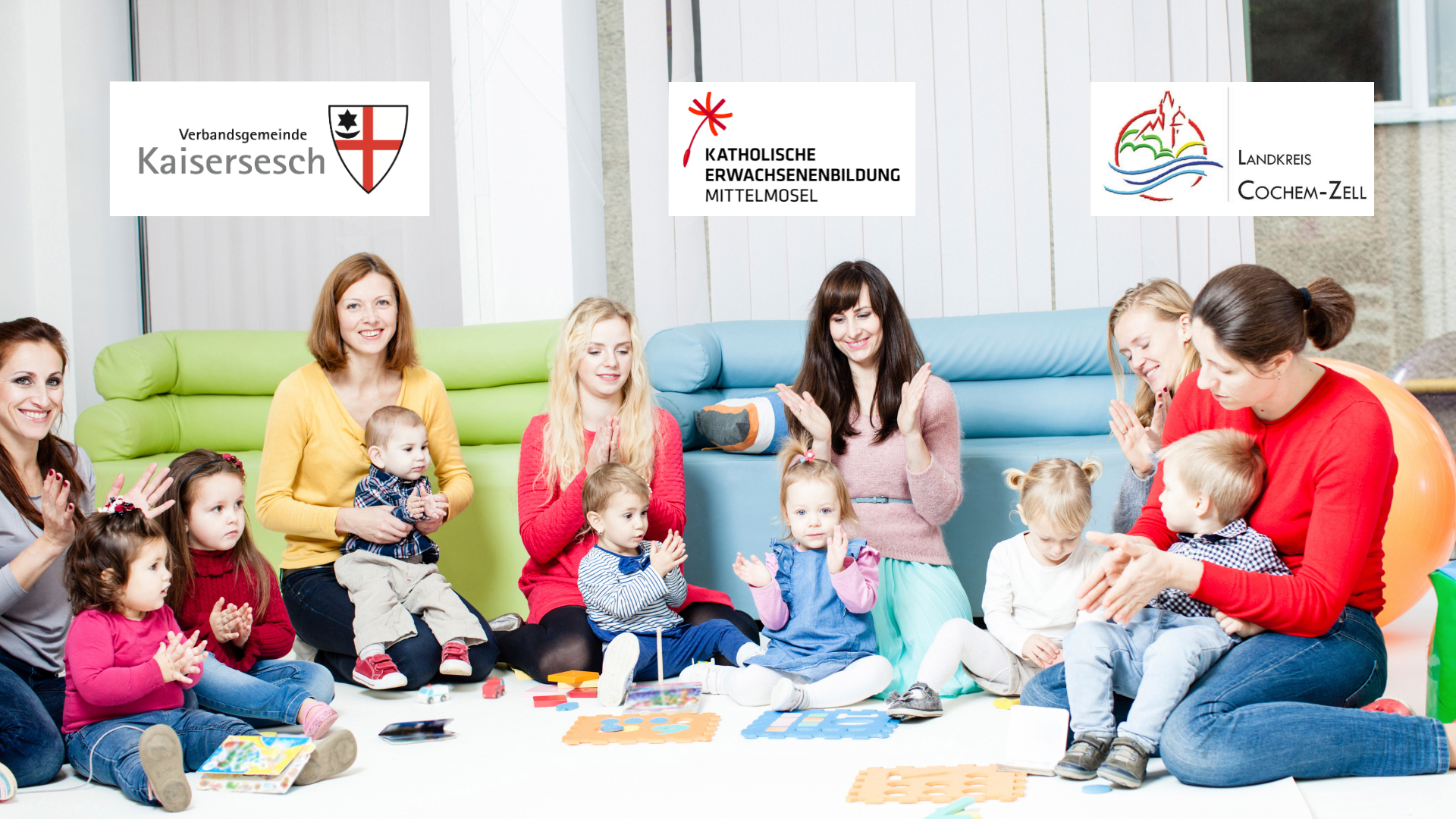 Das Bild zeigt eine Gruppe von Frauen und kleinen Kindern, die lächelnd und klatschend auf dem Boden sitzen. Im Hintergrund sind Sofas zu sehen. Oben im Bild sind drei Logos platziert: eines mit der Aufschrift „Verbandsgemeinde Kaisersesch“, ein weiteres mit „Katholische Erwachsenenbildung Mittelmosel“ und ein drittes mit „Landkreis Cochem-Zell“.  
