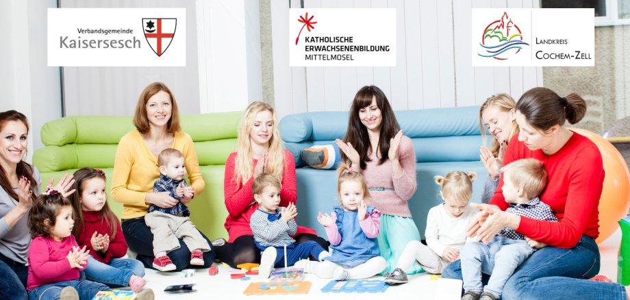 Mütter, die mit ihren Kindern spielen Das Bild zeigt eine Gruppe von Frauen und kleinen Kindern, die lächelnd und klatschend auf dem Boden sitzen. Im Hintergrund sind Sofas zu sehen. Oben im Bild sind drei Logos platziert: eines mit der Aufschrift „Verbandsgemeinde Kaisersesch“, ein weiteres mit „Katholische Erwachsenenbildung Mittelmosel“ und ein drittes mit „Landkreis Cochem-Zell“.