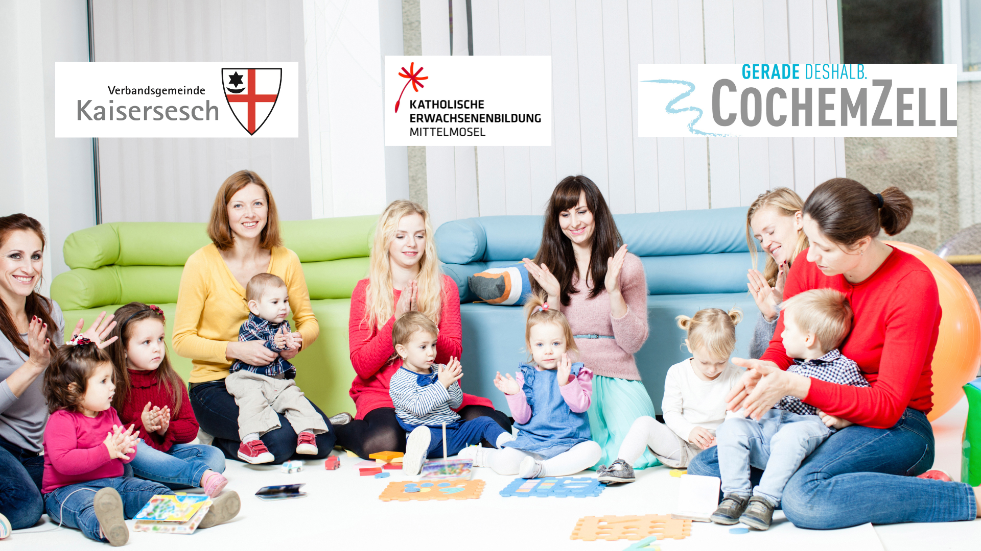 Das Bild zeigt eine Gruppe von Frauen und kleinen Kindern, die lächelnd und klatschend auf dem Boden sitzen. Im Hintergrund sind Sofas zu sehen. Oben im Bild sind drei Logos platziert: eines mit der Aufschrift „Verbandsgemeinde Kaisersesch“, ein weiteres mit „Katholische Erwachsenenbildung Mittelmosel“ und ein drittes mit „Gerade deshalb CochemZell“.  