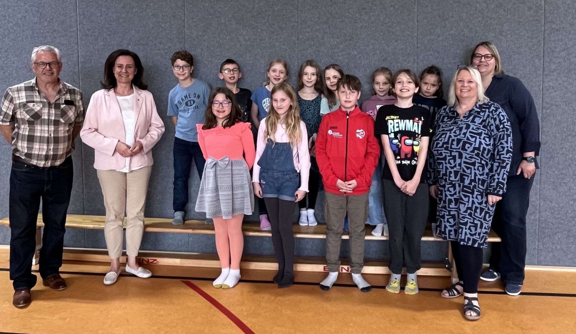 Das Bild zeigt eine Gruppe von 15 Personen, bestehend aus 12 Kindern und 3 Erwachsenen, die in einem Turnsaal auf einer Holzbühne stehen. Der Hintergrund ist eine graue Wand. Die Bühne ist aus Holz und hat mehrere Ebenen. Auf dem Boden des Turnsaals sind rote und gelbe Markierungslinien zu sehen. Von links nach rechts stehen zuerst Bürgermeister Albert Jung mit Brille und einem karierten Hemd und die rheinland-pfälzische Bildungsministerin Dr. Ute Eiling-Hütig mit einem hellrosa Blazer und einer weißen Bluse. Dahinter und daneben reihen sich die Kinder der Schulklasse 3a auf. Viele der Kinder sind mit hellen Farben bekleidet. Ein Kind trägt ein blaues T-Shirt mit der Aufschrift "GAME ON". Ein anderes Kind trägt eine rote Jacke. Einige Kinder tragen bunte T-Shirts mit Motiven. Die beiden erwachsenen Frauen ganz rechts im Bild (Schulleiterin Kathrin Matthias und Konrektorin Katja Dörrenbächer-Dietl) sind ebenfalls in der Gruppe. Eine davon trägt ein blaues, gemustertes Kleid und die andere eine dunkelblaue Jacke. Die meisten Kinder lächeln in die Kamera. Die gesamte Gruppe steht in einer lockeren Formation auf der Bühne. Die Beleuchtung ist hell und gleichmäßig. Der Fokus liegt auf den Personen, während die Umgebung leicht unscharf ist. Die Atmosphäre wirkt fröhlich und zusammengehörig.