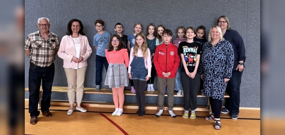 Das Bild zeigt eine Gruppe von 15 Personen, bestehend aus 12 Kindern und 3 Erwachsenen, die in einem Turnsaal auf einer Holzbühne stehen. Der Hintergrund ist eine graue Wand. Die Bühne ist aus Holz und hat mehrere Ebenen. Auf dem Boden des Turnsaals sind rote und gelbe Markierungslinien zu sehen. Von links nach rechts stehen zuerst Bürgermeister Albert Jung mit Brille und einem karierten Hemd und die rheinland-pfälzische Bildungsministerin Dr. Ute Eiling-Hütig mit einem hellrosa Blazer und einer weißen Bluse. Dahinter und daneben reihen sich die Kinder der Schulklasse 3a auf. Viele der Kinder sind mit hellen Farben bekleidet. Ein Kind trägt ein blaues T-Shirt mit der Aufschrift "GAME ON". Ein anderes Kind trägt eine rote Jacke. Einige Kinder tragen bunte T-Shirts mit Motiven. Die beiden erwachsenen Frauen ganz rechts im Bild (Schulleiterin Kathrin Matthias und Konrektorin Katja Dörrenbächer-Dietl) sind ebenfalls in der Gruppe. Eine davon trägt ein blaues, gemustertes Kleid und die andere eine dunkelblaue Jacke. Die meisten Kinder lächeln in die Kamera. Die gesamte Gruppe steht in einer lockeren Formation auf der Bühne. Die Beleuchtung ist hell und gleichmäßig. Der Fokus liegt auf den Personen, während die Umgebung leicht unscharf ist. Die Atmosphäre wirkt fröhlich und zusammengehörig.
