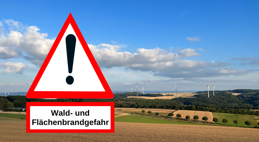 Das Bild zeigt eine Landschaftsaufnahme mit einem Warnschild im Vordergrund und einem weiten Blick auf Hügel und Windräder im Hintergrund. Im linken Vordergrund befindet sich ein großes, dreieckiges Warnschild mit einem roten Rand und weißem Hintergrund. In der Mitte des Dreiecks ist ein schwarzes Ausrufezeichen zu sehen. Unterhalb des Warnschilds befindet sich ein rechteckiges Schild mit weißem Hintergrund und rotem Rand, auf dem in schwarzer Schrift „Wald- und Flächenbrandgefahr“ steht. Diese beiden Schilder nehmen einen erheblichen Teil des linken Bildbereichs ein. Die Landschaft im Hintergrund ist in mehrere Ebenen unterteilt. Ganz vorn im Bild erstreckt sich ein Feld mit reifem, goldenem Getreide, das durch Ackerfurchen durchzogen ist. Dahinter folgt ein grüner Streifen, der wie eine Wiese oder ein anderes Feld aussieht. Weiter hinten erheben sich sanfte Hügel, die größtenteils mit dunklem Grün von Bäumen bedeckt sind. In der Ferne sind mehrere Windräder zu erkennen, die auf den Hügeln und Ebenen verteilt stehen. Einige Windräder sind weiter entfernt und scheinen kleiner, während andere näher sind und ihre Flügel sichtbar sind. Der Himmel ist ein leuchtendes Blau mit vereinzelten, flauschigen, weißen Wolken, die das Sonnenlicht reflektieren. Die Wolken sind in der oberen Hälfte des Bildes verteilt und verleihen dem Himmel Tiefe. Die Farben im Bild sind kräftig und natürlich. Das Gold des Getreides, das intensive Grün der Bäume und Felder, das Blau des Himmels und das Weiß der Wolken bilden einen harmonischen Kontrast. Die roten Ränder der Schilder heben sich deutlich ab und lenken die Aufmerksamkeit auf die Warnung. Die Komposition des Bildes ist auf das Warnschild fokussiert, das prominent im linken Drittel platziert ist. Die Landschaft im Hintergrund dient als malerischer Kontext, der die potenzielle Gefahr von Wald- und Flächenbränden unterstreicht. Die Linien der Felder und Hügel führen das Auge des Betrachters in die Tiefe des Bildes.