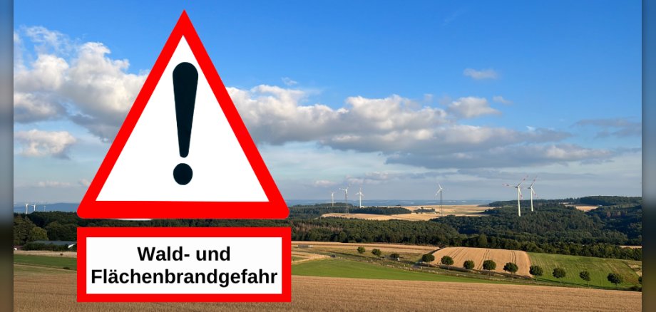 Das Bild zeigt eine Landschaftsaufnahme mit einem Warnschild im Vordergrund und einem weiten Blick auf Hügel und Windräder im Hintergrund. Im linken Vordergrund befindet sich ein großes, dreieckiges Warnschild mit einem roten Rand und weißem Hintergrund. In der Mitte des Dreiecks ist ein schwarzes Ausrufezeichen zu sehen. Unterhalb des Warnschilds befindet sich ein rechteckiges Schild mit weißem Hintergrund und rotem Rand, auf dem in schwarzer Schrift „Wald- und Flächenbrandgefahr“ steht. Diese beiden Schilder nehmen einen erheblichen Teil des linken Bildbereichs ein. Die Landschaft im Hintergrund ist in mehrere Ebenen unterteilt. Ganz vorn im Bild erstreckt sich ein Feld mit reifem, goldenem Getreide, das durch Ackerfurchen durchzogen ist. Dahinter folgt ein grüner Streifen, der wie eine Wiese oder ein anderes Feld aussieht. Weiter hinten erheben sich sanfte Hügel, die größtenteils mit dunklem Grün von Bäumen bedeckt sind. In der Ferne sind mehrere Windräder zu erkennen, die auf den Hügeln und Ebenen verteilt stehen. Einige Windräder sind weiter entfernt und scheinen kleiner, während andere näher sind und ihre Flügel sichtbar sind. Der Himmel ist ein leuchtendes Blau mit vereinzelten, flauschigen, weißen Wolken, die das Sonnenlicht reflektieren. Die Wolken sind in der oberen Hälfte des Bildes verteilt und verleihen dem Himmel Tiefe. Die Farben im Bild sind kräftig und natürlich. Das Gold des Getreides, das intensive Grün der Bäume und Felder, das Blau des Himmels und das Weiß der Wolken bilden einen harmonischen Kontrast. Die roten Ränder der Schilder heben sich deutlich ab und lenken die Aufmerksamkeit auf die Warnung. Die Komposition des Bildes ist auf das Warnschild fokussiert, das prominent im linken Drittel platziert ist. Die Landschaft im Hintergrund dient als malerischer Kontext, der die potenzielle Gefahr von Wald- und Flächenbränden unterstreicht. Die Linien der Felder und Hügel führen das Auge des Betrachters in die Tiefe des Bildes.