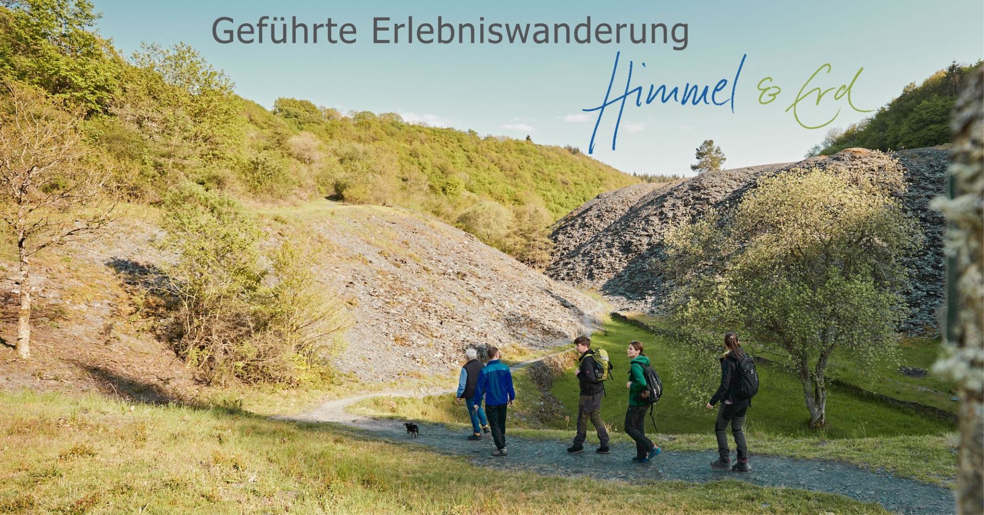 Das Bild zeigt eine Gruppe von vier Wanderern und einen kleinen schwarzen Hund, die auf einem schmalen, gewundenen Weg durch eine hügelige, grüne Landschaft gehen. Die Szene ist von oben beleuchtet, was auf einen sonnigen Tag hindeutet. Die Wanderer sind von links nach rechts: ein älterer Mann mit einer blauen Weste über einem Hemd und Jeans, gefolgt von einem jüngeren Mann in einer blauen Jacke, einer dunklen Hose und Schuhen. Dann kommt ein Mann mit einem grünen Rucksack auf dem Rücken, einer dunklen Jacke und grauen Hosen. Zuletzt geht eine Frau mit einem schwarzen Rucksack, einer dunklen Hose und einem grünen Oberteil. Der kleine schwarze Hund läuft auf dem Weg vor der Gruppe. Der Weg schlängelt sich durch eine von der Sonne beschienene Senke. Links des Weges erhebt sich ein steiler, felsiger Hang, der von niedriger Vegetation und einigen kahlen Bäumen bedeckt ist. Die Felsen sind hellbraun und grau. Rechts des Weges sind weitere felsige Hänge zu sehen, die mit schattigen Bereichen durchsetzt sind. Weiter oben auf den Hügeln sind dichte, grüne Wälder zu erkennen, die in einem hellen Himmel verschwimmen. Die Farben im Bild sind überwiegend natürliche Töne: viele Grüntöne von der Vegetation, verschiedene Brauntöne und Grau von den Felsen und dem Weg, sowie das klare Blau des Himmels. Das Licht spielt eine wichtige Rolle und wirft Schatten auf den Boden und die Hänge, was der Szene Tiefe verleiht. Oben rechts im Bild sind in einer geschwungenen, blauen und grünen Schrift die Worte "Himmel & Erd" zu sehen. Über diesem Schriftzug steht in größerer, grauer Schrift "Geführte Erlebniswanderung". Die Komposition ist so angeordnet, dass die Wanderer den Weg entlanggehen, was eine Bewegung und Richtung in das Bild bringt. Die Landschaft mit den Hängen und Wäldern rahmt die Gruppe ein und betont die Weite und Schönheit der Natur. Der Vordergrund ist grün und leicht unscharf, was die Wanderer und den Weg in den Fokus rückt. Der Himmel nimmt einen beträchtlichen Teil des oberen Bildbereichs ein und erzeugt ein Gefühl von Offenheit.