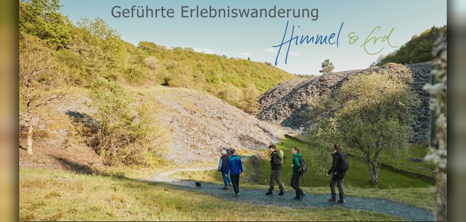 Das Bild zeigt eine Gruppe von vier Wanderern und einen kleinen schwarzen Hund, die auf einem schmalen, gewundenen Weg durch eine hügelige, grüne Landschaft gehen. Die Szene ist von oben beleuchtet, was auf einen sonnigen Tag hindeutet. Die Wanderer sind von links nach rechts: ein älterer Mann mit einer blauen Weste über einem Hemd und Jeans, gefolgt von einem jüngeren Mann in einer blauen Jacke, einer dunklen Hose und Schuhen. Dann kommt ein Mann mit einem grünen Rucksack auf dem Rücken, einer dunklen Jacke und grauen Hosen. Zuletzt geht eine Frau mit einem schwarzen Rucksack, einer dunklen Hose und einem grünen Oberteil. Der kleine schwarze Hund läuft auf dem Weg vor der Gruppe. Der Weg schlängelt sich durch eine von der Sonne beschienene Senke. Links des Weges erhebt sich ein steiler, felsiger Hang, der von niedriger Vegetation und einigen kahlen Bäumen bedeckt ist. Die Felsen sind hellbraun und grau. Rechts des Weges sind weitere felsige Hänge zu sehen, die mit schattigen Bereichen durchsetzt sind. Weiter oben auf den Hügeln sind dichte, grüne Wälder zu erkennen, die in einem hellen Himmel verschwimmen. Die Farben im Bild sind überwiegend natürliche Töne: viele Grüntöne von der Vegetation, verschiedene Brauntöne und Grau von den Felsen und dem Weg, sowie das klare Blau des Himmels. Das Licht spielt eine wichtige Rolle und wirft Schatten auf den Boden und die Hänge, was der Szene Tiefe verleiht. Oben rechts im Bild sind in einer geschwungenen, blauen und grünen Schrift die Worte "Himmel & Erd" zu sehen. Über diesem Schriftzug steht in größerer, grauer Schrift "Geführte Erlebniswanderung". Die Komposition ist so angeordnet, dass die Wanderer den Weg entlanggehen, was eine Bewegung und Richtung in das Bild bringt. Die Landschaft mit den Hängen und Wäldern rahmt die Gruppe ein und betont die Weite und Schönheit der Natur. Der Vordergrund ist grün und leicht unscharf, was die Wanderer und den Weg in den Fokus rückt. Der Himmel nimmt einen beträchtlichen Teil des oberen Bildbereichs ein und erzeugt ein Gefühl von Offenheit.