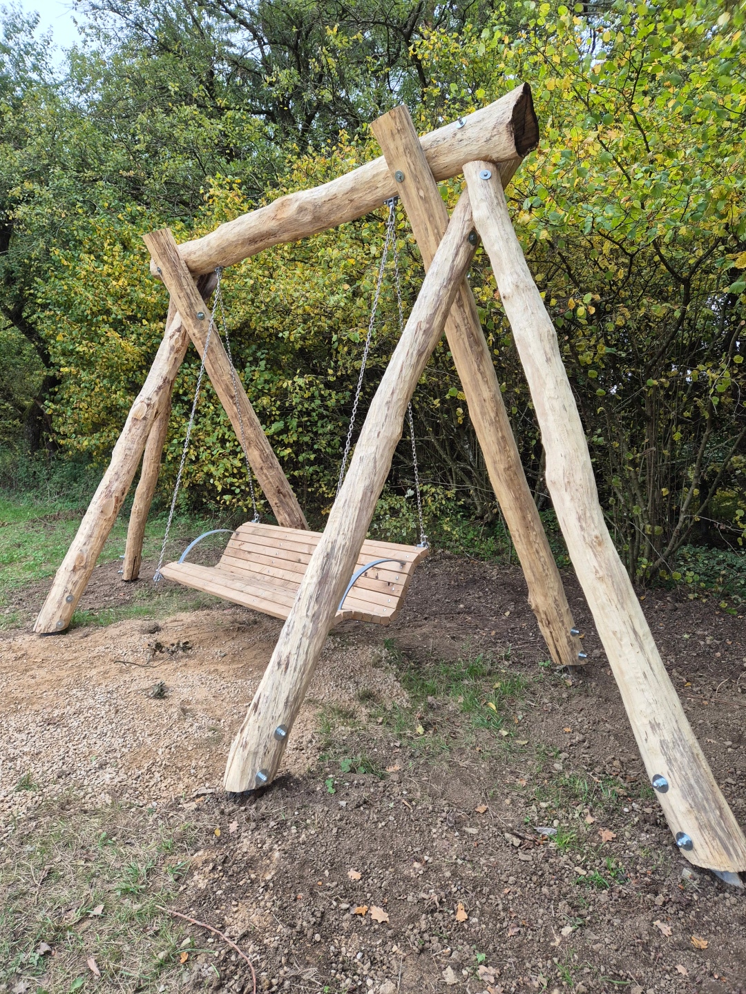 Das Bild zeigt eine robuste Schaukel aus groben, unbehandelten Holzstämmen. Die Schaukelkonstruktion besteht aus vier schräg aufgestellten Stämmen, die oben mit einem waagerechten Balken verbunden sind. Die Holzoberflächen sind rau und zeigen Astlöcher und Rindenreste, was dem Ganzen einen rustikalen und natürlichen Look verleiht. Große Schrauben mit Muttern verbinden die einzelnen Holzteile. Die Sitzfläche der Schaukel ist aus mehreren Holzplanken gefertigt und hängt an zwei stabilen Metallketten. Die Sitzfläche selbst ist hellbraun und wirkt ebenfalls massiv. Die Schaukel ist im Freien aufgestellt, der Boden besteht aus einer Mischung aus Sand, Kies und Erde, mit einigen Grashalmen und Herbstblättern darauf. Im Hintergrund ist dichter Baumbestand mit grünen und gelblichen Blättern zu sehen, was auf eine herbstliche Jahreszeit hindeutet. Die Beleuchtung ist diffus, was auf einen bewölkten Himmel oder eine Zeit ohne direkte Sonneneinstrahlung schließen lässt. Die Komposition ist zentriert auf die Schaukel, die fast den gesamten unteren und mittleren Bildbereich einnimmt. Die natürliche Umgebung und das grobe Holz schaffen eine Atmosphäre der Ruhe und Gemütlichkeit.