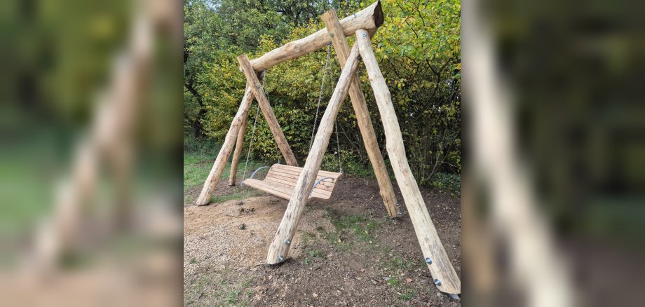 Eine Schaukel aus unbehandelten Baumstämmen und einer Holzbank hängt an stabilen Metallketten. Die Schaukel ist im Freien auf einem Kies- und Erdboden aufgestellt, umgeben von grünem und gelb gefärbtem Laub. Das Bild zeigt eine robuste Schaukel aus groben, unbehandelten Holzstämmen. Die Schaukelkonstruktion besteht aus vier schräg aufgestellten Stämmen, die oben mit einem waagerechten Balken verbunden sind. Die Holzoberflächen sind rau und zeigen Astlöcher und Rindenreste, was dem Ganzen einen rustikalen und natürlichen Look verleiht. Große Schrauben mit Muttern verbinden die einzelnen Holzteile. Die Sitzfläche der Schaukel ist aus mehreren Holzplanken gefertigt und hängt an zwei stabilen Metallketten. Die Sitzfläche selbst ist hellbraun und wirkt ebenfalls massiv. Die Schaukel ist im Freien aufgestellt, der Boden besteht aus einer Mischung aus Sand, Kies und Erde, mit einigen Grashalmen und Herbstblättern darauf. Im Hintergrund ist dichter Baumbestand mit grünen und gelblichen Blättern zu sehen, was auf eine herbstliche Jahreszeit hindeutet. Die Beleuchtung ist diffus, was auf einen bewölkten Himmel oder eine Zeit ohne direkte Sonneneinstrahlung schließen lässt. Die Komposition ist zentriert auf die Schaukel, die fast den gesamten unteren und mittleren Bildbereich einnimmt. Die natürliche Umgebung und das grobe Holz schaffen eine Atmosphäre der Ruhe und Gemütlichkeit.