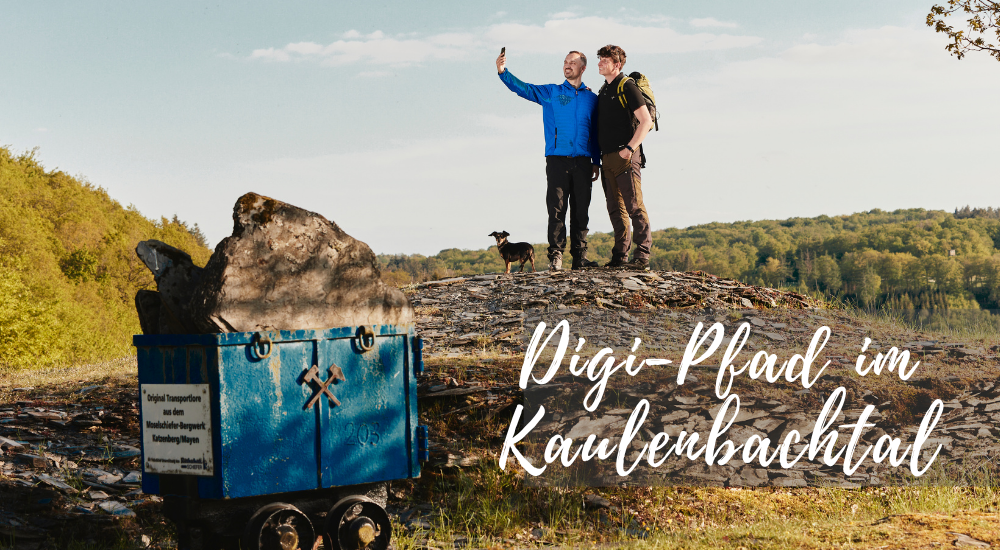 Das Bild zeigt eine weite Landschaft, das Kaulenbachtal, unter einem hellblauen Himmel mit vereinzelten weißen Wolken. Im Vordergrund links befindet sich ein kleiner, blau angestrichener Minenwagen, der mit einem großen grauen Stein beladen ist. Auf dem Wagen ist ein Schild mit deutscher Schrift und einem gekreuzten Hammer- und Schlegel-Symbol angebracht. Der Wagen steht auf unebenem, felsigem Boden, der teilweise von grünem Gras bedeckt ist. Im mittleren Bereich des Bildes stehen zwei Männer nebeneinander auf einer Anhöhe aus Steinen und Erde. Der Mann links trägt eine blaue Jacke und hält ein Smartphone in der Hand, um ein Selfie zu machen. Er lächelt in die Kamera. Der Mann rechts trägt ein schwarzes T-Shirt, eine grüne Weste mit Rucksack und schaut ebenfalls in die Richtung des Smartphones. Beide Männer tragen Wanderausrüstung. Ein kleiner, dunkler Hund steht zwischen den beiden Männern und blickt in die gleiche Richtung. Im Hintergrund erstreckt sich eine dicht bewaldete Hügellandschaft. Die Bäume sind in verschiedenen Grüntönen gehalten und deuten auf einen Frühlingstag oder frühen Sommer hin. Die Sonne scheint von rechts, was leichte Schatten auf den Boden und die Felsen wirft. Über den Köpfen der Männer und im rechten Teil des Bildes sind in geschwungener, weißer Schrift die Worte "Digi-Pfad im Kaubenbachtal" zu lesen. Die Farbpalette des Bildes ist von natürlichen Tönen geprägt: Blau des Wagens und des Himmels, Grau des Steins und der Felsen, verschiedene Grüntöne der Vegetation, Braun der Kleidung und des Hundes sowie Weiß der Schrift und der Wolken. Die Komposition ist ausgewogen, wobei der Minenwagen und die beiden Männer mit dem Hund die zentralen Elemente bilden. Der weite Hintergrund mit der bewaldeten Landschaft verleiht dem Bild Tiefe und Weite. Die Aufnahme scheint mit einer leichten Weitwinkeloptik gemacht worden zu sein, um die Landschaft des Kaulenbachtals umfassend darzustellen. Das Licht ist hell und natürlich, was eine angenehme und lebendige Atmosphäre schafft.