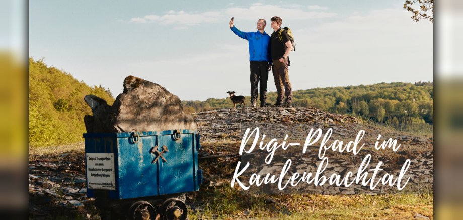 Das Bild zeigt eine weite Landschaft, das Kaulenbachtal, unter einem hellblauen Himmel mit vereinzelten weißen Wolken. Im Vordergrund links befindet sich ein kleiner, blau angestrichener Minenwagen, der mit einem großen grauen Stein beladen ist. Auf dem Wagen ist ein Schild mit deutscher Schrift und einem gekreuzten Hammer- und Schlegel-Symbol angebracht. Der Wagen steht auf unebenem, felsigem Boden, der teilweise von grünem Gras bedeckt ist. Im mittleren Bereich des Bildes stehen zwei Männer nebeneinander auf einer Anhöhe aus Steinen und Erde. Der Mann links trägt eine blaue Jacke und hält ein Smartphone in der Hand, um ein Selfie zu machen. Er lächelt in die Kamera. Der Mann rechts trägt ein schwarzes T-Shirt, eine grüne Weste mit Rucksack und schaut ebenfalls in die Richtung des Smartphones. Beide Männer tragen Wanderausrüstung. Ein kleiner, dunkler Hund steht zwischen den beiden Männern und blickt in die gleiche Richtung. Im Hintergrund erstreckt sich eine dicht bewaldete Hügellandschaft. Die Bäume sind in verschiedenen Grüntönen gehalten und deuten auf einen Frühlingstag oder frühen Sommer hin. Die Sonne scheint von rechts, was leichte Schatten auf den Boden und die Felsen wirft. Über den Köpfen der Männer und im rechten Teil des Bildes sind in geschwungener, weißer Schrift die Worte "Digi-Pfad im Kaubenbachtal" zu lesen. Die Farbpalette des Bildes ist von natürlichen Tönen geprägt: Blau des Wagens und des Himmels, Grau des Steins und der Felsen, verschiedene Grüntöne der Vegetation, Braun der Kleidung und des Hundes sowie Weiß der Schrift und der Wolken. Die Komposition ist ausgewogen, wobei der Minenwagen und die beiden Männer mit dem Hund die zentralen Elemente bilden. Der weite Hintergrund mit der bewaldeten Landschaft verleiht dem Bild Tiefe und Weite. Die Aufnahme scheint mit einer leichten Weitwinkeloptik gemacht worden zu sein, um die Landschaft des Kaulenbachtals umfassend darzustellen. Das Licht ist hell und natürlich, was eine angenehme und lebendige Atmosphäre schafft.