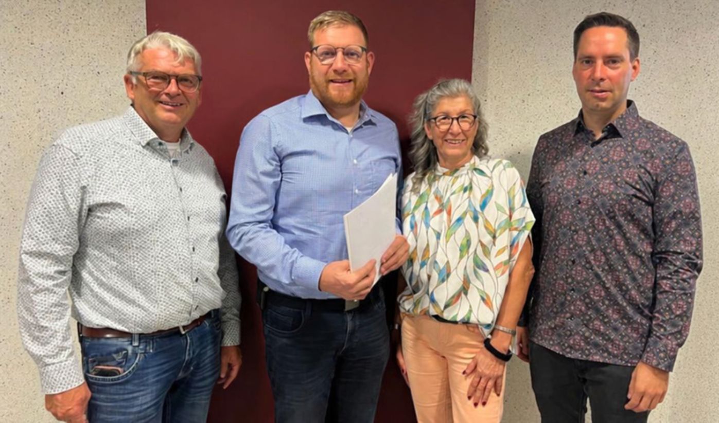 Gruppenfoto anlässlich des 25‑jährigen Dienstjubiläums von Daniel Hermes. Auf dem Foto sind vier Personen zu sehen, davon drei Männer und eine Frau. Die Personen sind bunt angezogen und lächeln. Der Hintergrund ist dunkelrot und hellbeige. 