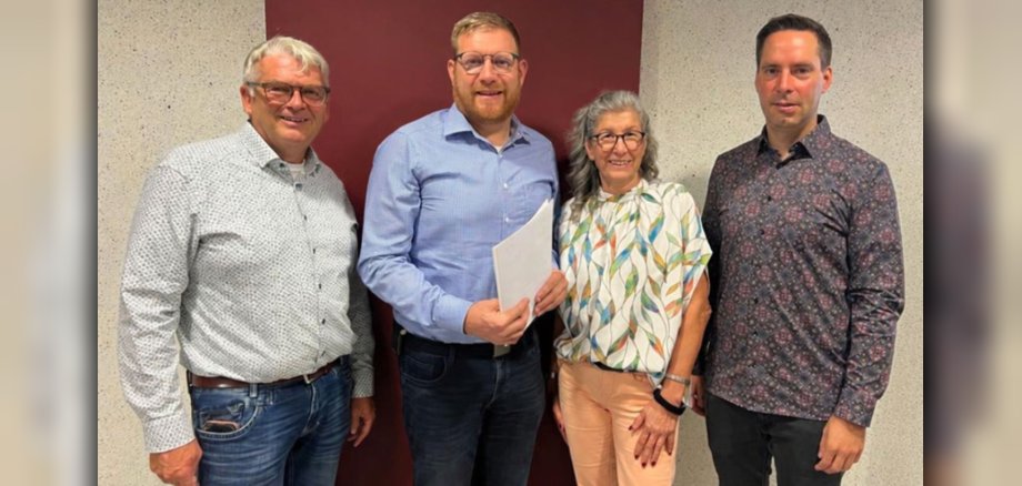 Gruppenfoto anlässlich des 25‑jährigen Dienstjubiläums von Daniel Hermes. Gruppenfoto anlässlich des 25‑jährigen Dienstjubiläums von Daniel Hermes. Auf dem Foto sind vier Personen zu sehen, davon drei Männer und eine Frau. Die Personen sind bunt angezogen und lächeln. Der Hintergrund ist dunkelrot und hellbeige.