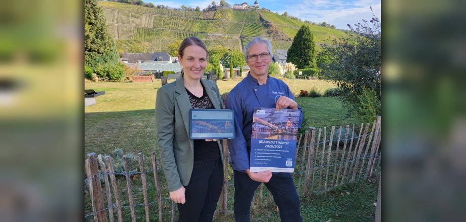 Rudi Burch und Steffi Schmitz vom Hotel „Zur Marienburg“ organisieren das neue „Winterwunderland“ auf der Terrasse und im Garten des Hotels. Eine Frau und ein Mann präsentieren ein Tablet und einen Flyer zur „(R)Auszeit Winter 2026/2027“ vor einer sonnigen Weinberglandschaft.