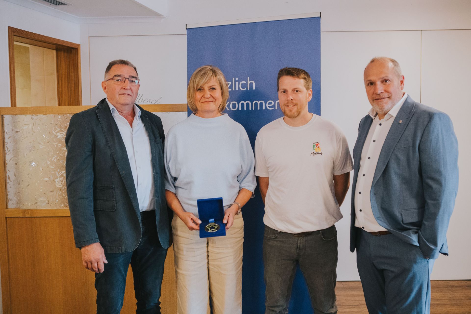 Vier Erwachsene stehen nebeneinander in einem Raum; die Frau in der Mitte hält eine geöffnete blaue Schatulle mit einer Medaille. Im Hintergrund steht ein blaues Banner.
