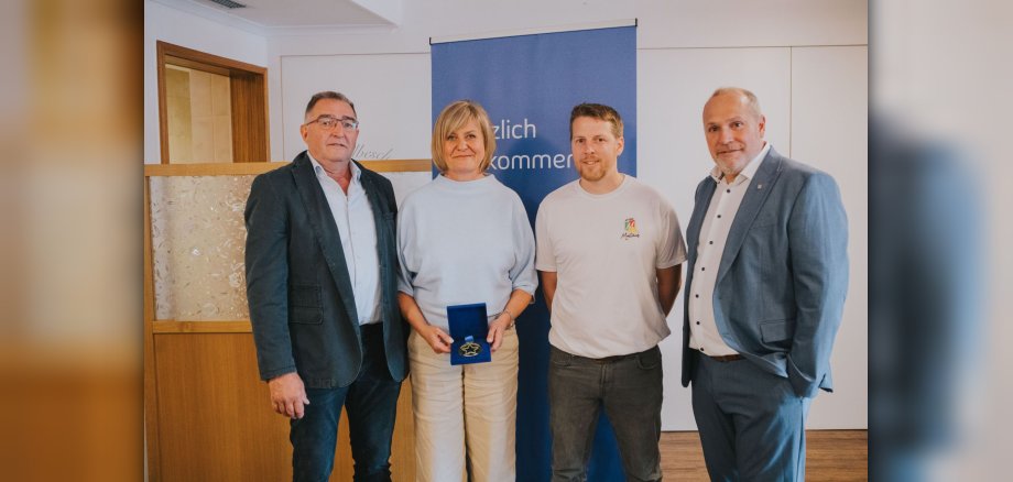 V. l. n. r.: Beigeordneter Thomas Welter, Tanja Knieper (Vorstandsmitglied) und Peter Gilles (1. Vorsitzender) vom Verein zur Förderung der Dorfkultur sowie Thomas Hau (Kommunalmanager von Westenergie) Vier Erwachsene stehen nebeneinander in einem Raum; die Frau in der Mitte hält eine geöffnete blaue Schatulle mit einer Medaille. Im Hintergrund steht ein blaues Banner.