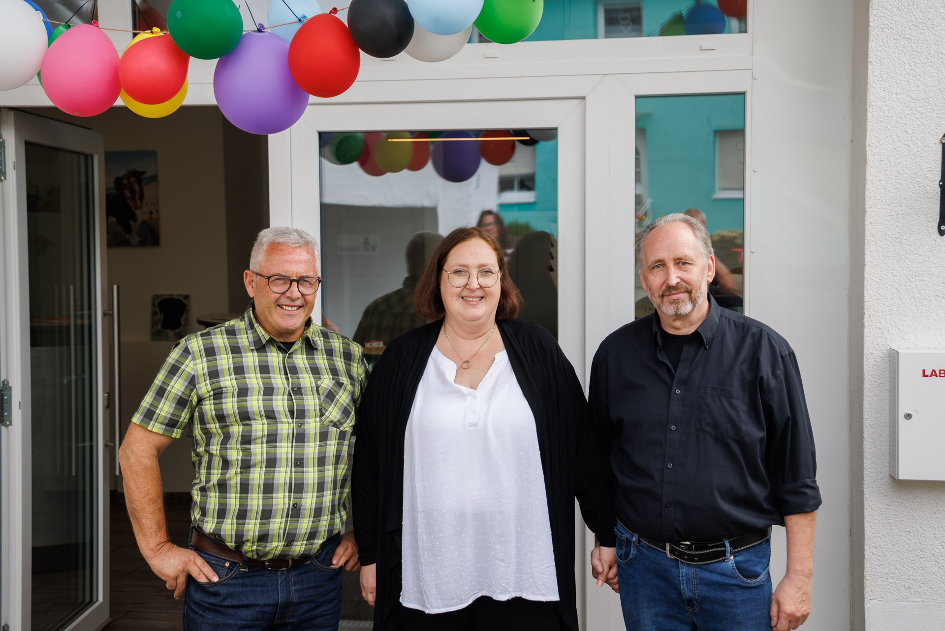 Drei Personen stehen lächelnd vor einem Hauseingang, der mit bunten Luftballons geschmückt ist.