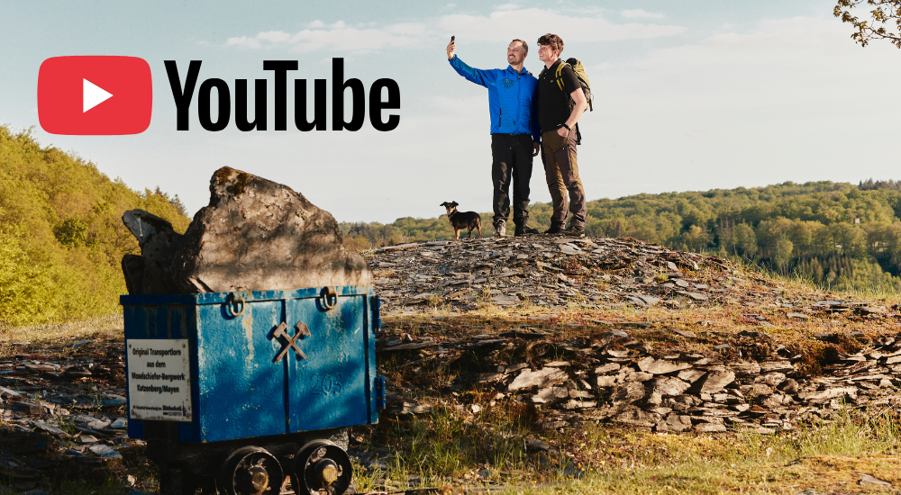 Zwei Wanderer mit Hund stehen auf einer Halde neben einem alten Förderwagen; oben links ist das YouTube-Logo zu sehen.