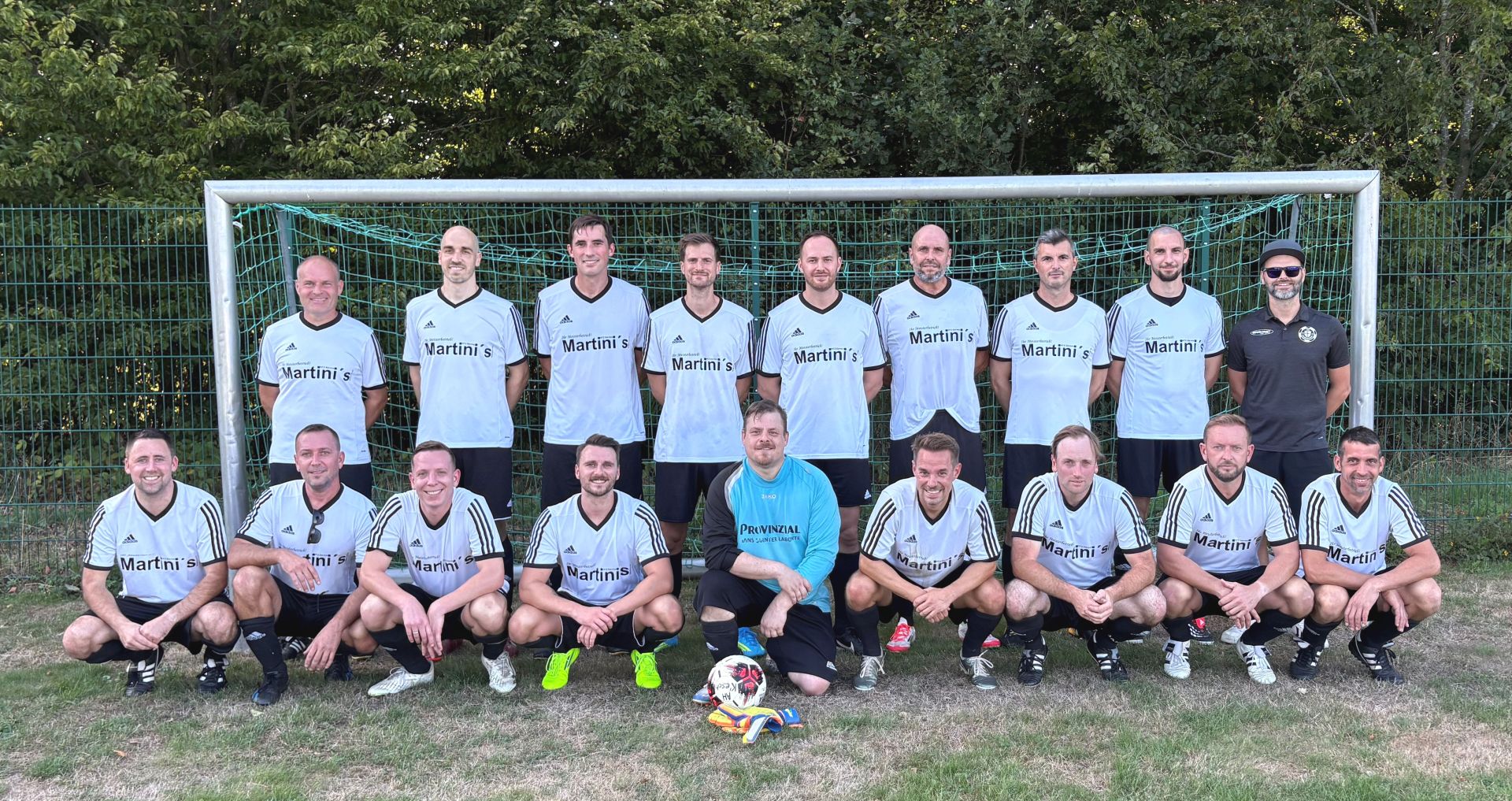 Fußballmannschaft in weißen Trikots posiert gemeinsam mit einem Torwart im blauen Trikot und einem Trainer vor einem Fußballtor; vor der Gruppe liegt ein Fußball auf dem Rasen.