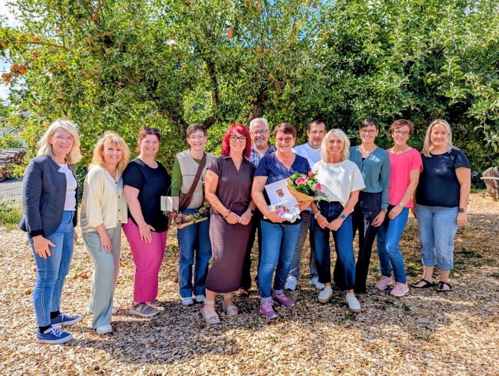 Eine Gruppe von zwölf Personen steht bei sonnigem Wetter vor einem Obstbaum und lächelt in die Kamera. In der Mitte hält eine Person Blumen und Geschenke.