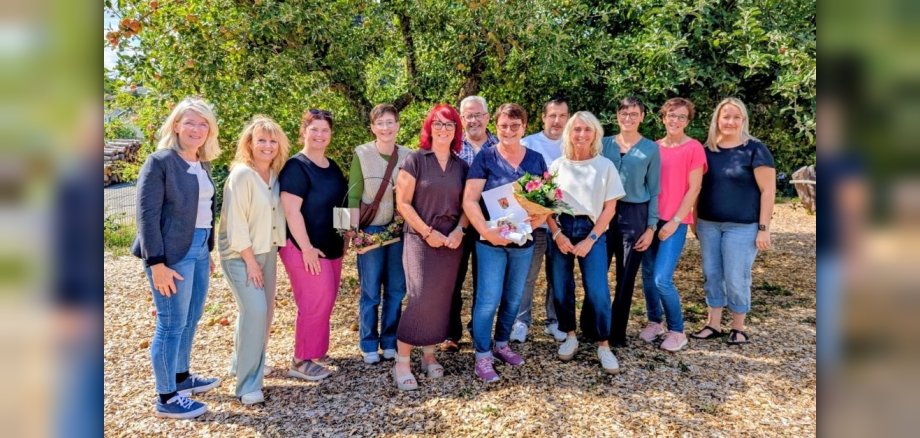 Eine Gruppe von zwölf Personen steht bei sonnigem Wetter vor einem Obstbaum und lächelt in die Kamera. In der Mitte hält eine Person Blumen und Geschenke.