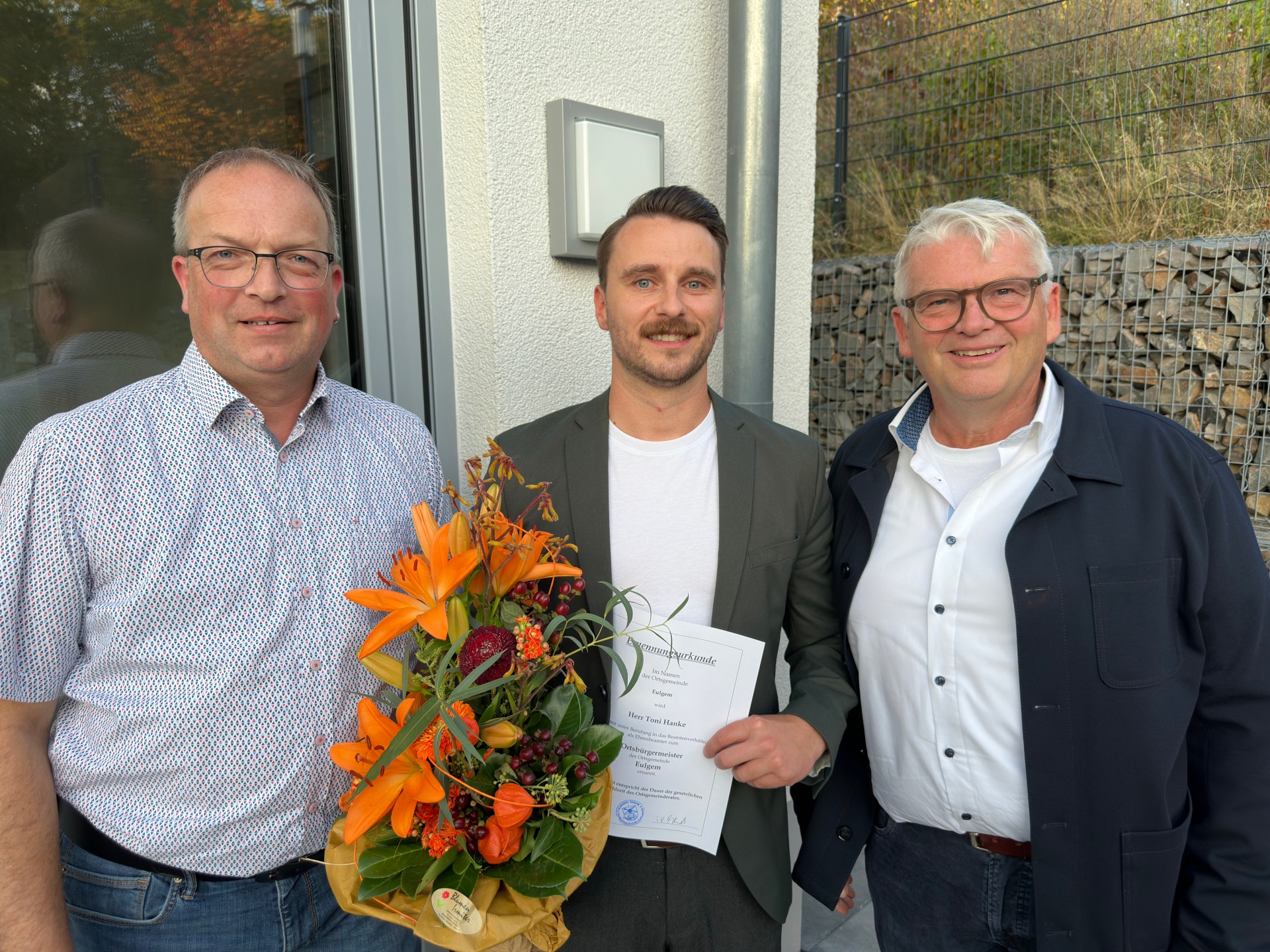 Drei Männer stehen zusammen vor einem Gebäude. Einer der Männer hält eine Urkunde und einen Blumenstrauß in der Hand. 