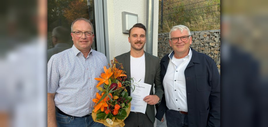 Toni Hanke ist neuer Ortsbürgermeister von Eulgem Drei Männer stehen zusammen vor einem Gebäude. Einer der Männer hält eine Urkunde und einen Blumenstrauß in der Hand.