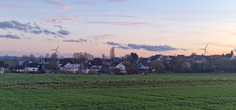 Das Bild zeigt die Ortsgemeinde Zettingen in der Dämmerung oder bei Sonnenuntergang. Im Vordergrund erstreckt sich ein grünes Feld. Dahinter ist eine Ansammlung von Häusern zu sehen, die Zettingen zeigen, umgeben von Bäumen, die größtenteils kahl sind. Am Horizont, über den Dächern, ragen zwei Windräder in den Himmel. Auf der rechten Seite ist auch ein Kirchturm erkennbar. Der Himmel ist in sanften Pastelltönen von Rosa, Orange und Hellblau gehalten, mit einigen verstreuten Wolken.